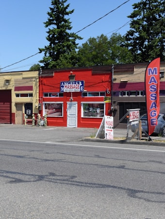 A small massage parlor with a bright red facade is located along a street. It features a prominent blue and white sign with the word 'MASSAGE' above the entrance. There is a large red vertical banner with the same word on the sidewalk. Several potted plants and decorations are placed near the door.