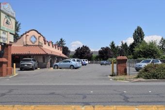 A Mexican-style restaurant with a red tile roof is situated next to a parking lot with several cars parked. The sign outside displays the restaurant's name and mentions margaritas to go. Surrounding the area are trees and bushes with a clear blue sky in the background.