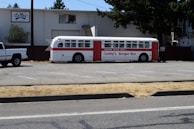 A vintage bus painted white with red accents and a sign that reads 'Lucky's Burger Bus' is parked in a nearly empty parking lot next to a white pickup truck. The surroundings include a warehouse-like building with a sign that says LeMay, some greenery, and trees providing partial shade.