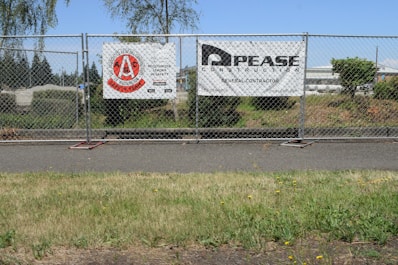 A chain-link fence with two signs attached stands along a sidewalk. The left sign features a red circular logo with text indicating a construction safety team. The right sign displays the name and information of a general contractor. Behind the fence, there is a grassy area with bushes and trees, and an industrial area with buildings in the background under a clear blue sky.