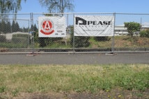 A chain-link fence with two signs attached stands along a sidewalk. The left sign features a red circular logo with text indicating a construction safety team. The right sign displays the name and information of a general contractor. Behind the fence, there is a grassy area with bushes and trees, and an industrial area with buildings in the background under a clear blue sky.