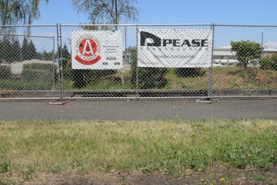 A chain-link fence with two signs attached stands along a sidewalk. The left sign features a red circular logo with text indicating a construction safety team. The right sign displays the name and information of a general contractor. Behind the fence, there is a grassy area with bushes and trees, and an industrial area with buildings in the background under a clear blue sky.