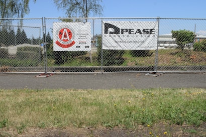 A chain-link fence with two signs attached stands along a sidewalk. The left sign features a red circular logo with text indicating a construction safety team. The right sign displays the name and information of a general contractor. Behind the fence, there is a grassy area with bushes and trees, and an industrial area with buildings in the background under a clear blue sky.