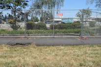A chain-link fence is set up along a sidewalk with a 'Danger Construction Area Keep Out' sign attached. Beyond the fence, there are trees, shrubs, and a large blue shipping container in a parking lot. The sky is clear and blue, and the area appears to be part of a construction or development site.