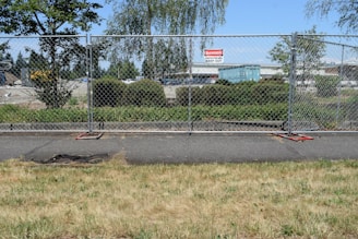 A chain-link fence is set up along a sidewalk with a 'Danger Construction Area Keep Out' sign attached. Beyond the fence, there are trees, shrubs, and a large blue shipping container in a parking lot. The sky is clear and blue, and the area appears to be part of a construction or development site.