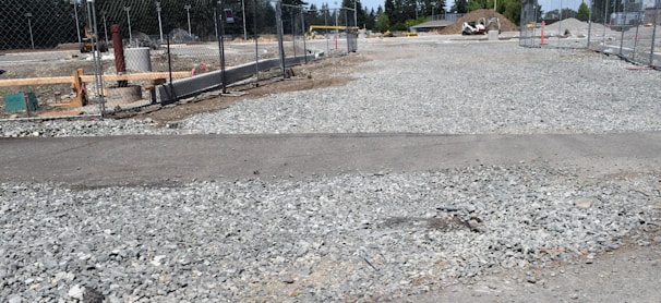 A construction site with gravel spread across the ground, surrounded by a chain-link fence. Several construction-related objects like pipes and building materials are visible. In the background, there is a mound of dirt and some trees.