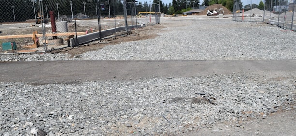 A construction site with gravel spread across the ground, surrounded by a chain-link fence. Several construction-related objects like pipes and building materials are visible. In the background, there is a mound of dirt and some trees.