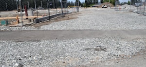 A construction site with gravel spread across the ground, surrounded by a chain-link fence. Several construction-related objects like pipes and building materials are visible. In the background, there is a mound of dirt and some trees.