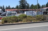 A fenced commercial area with vehicles and stacked materials, such as pallets, visible inside. The setting includes a parking lot and storage area, with a backdrop of tall trees. The fence has a black metal design, and a layer of greenery lines the outer edge of the premises.