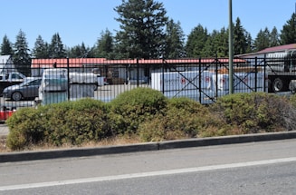 A fenced commercial area with vehicles and stacked materials, such as pallets, visible inside. The setting includes a parking lot and storage area, with a backdrop of tall trees. The fence has a black metal design, and a layer of greenery lines the outer edge of the premises.