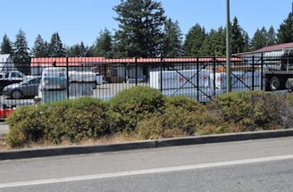 A fenced commercial area with vehicles and stacked materials, such as pallets, visible inside. The setting includes a parking lot and storage area, with a backdrop of tall trees. The fence has a black metal design, and a layer of greenery lines the outer edge of the premises.