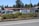A fenced commercial area with vehicles and stacked materials, such as pallets, visible inside. The setting includes a parking lot and storage area, with a backdrop of tall trees. The fence has a black metal design, and a layer of greenery lines the outer edge of the premises.