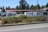 A fenced commercial area with vehicles and stacked materials, such as pallets, visible inside. The setting includes a parking lot and storage area, with a backdrop of tall trees. The fence has a black metal design, and a layer of greenery lines the outer edge of the premises.