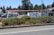 A fenced commercial area with vehicles and stacked materials, such as pallets, visible inside. The setting includes a parking lot and storage area, with a backdrop of tall trees. The fence has a black metal design, and a layer of greenery lines the outer edge of the premises.