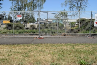 A chain-link fence with signs warning of a construction site surrounds an area with bushes and trees. Behind the fence, construction equipment and vehicles are visible, indicating work in progress. The scene also includes a paved pathway and grass in the foreground.