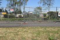A chain-link fence with signs warning of a construction site surrounds an area with bushes and trees. Behind the fence, construction equipment and vehicles are visible, indicating work in progress. The scene also includes a paved pathway and grass in the foreground.