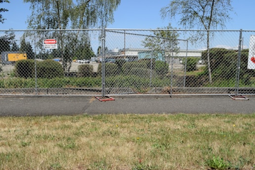 A chain-link fence with signs warning of a construction site surrounds an area with bushes and trees. Behind the fence, construction equipment and vehicles are visible, indicating work in progress. The scene also includes a paved pathway and grass in the foreground.