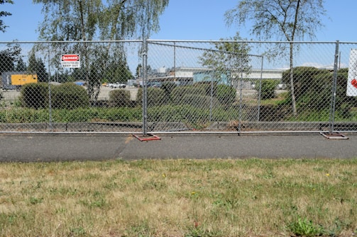 A chain-link fence with signs warning of a construction site surrounds an area with bushes and trees. Behind the fence, construction equipment and vehicles are visible, indicating work in progress. The scene also includes a paved pathway and grass in the foreground.