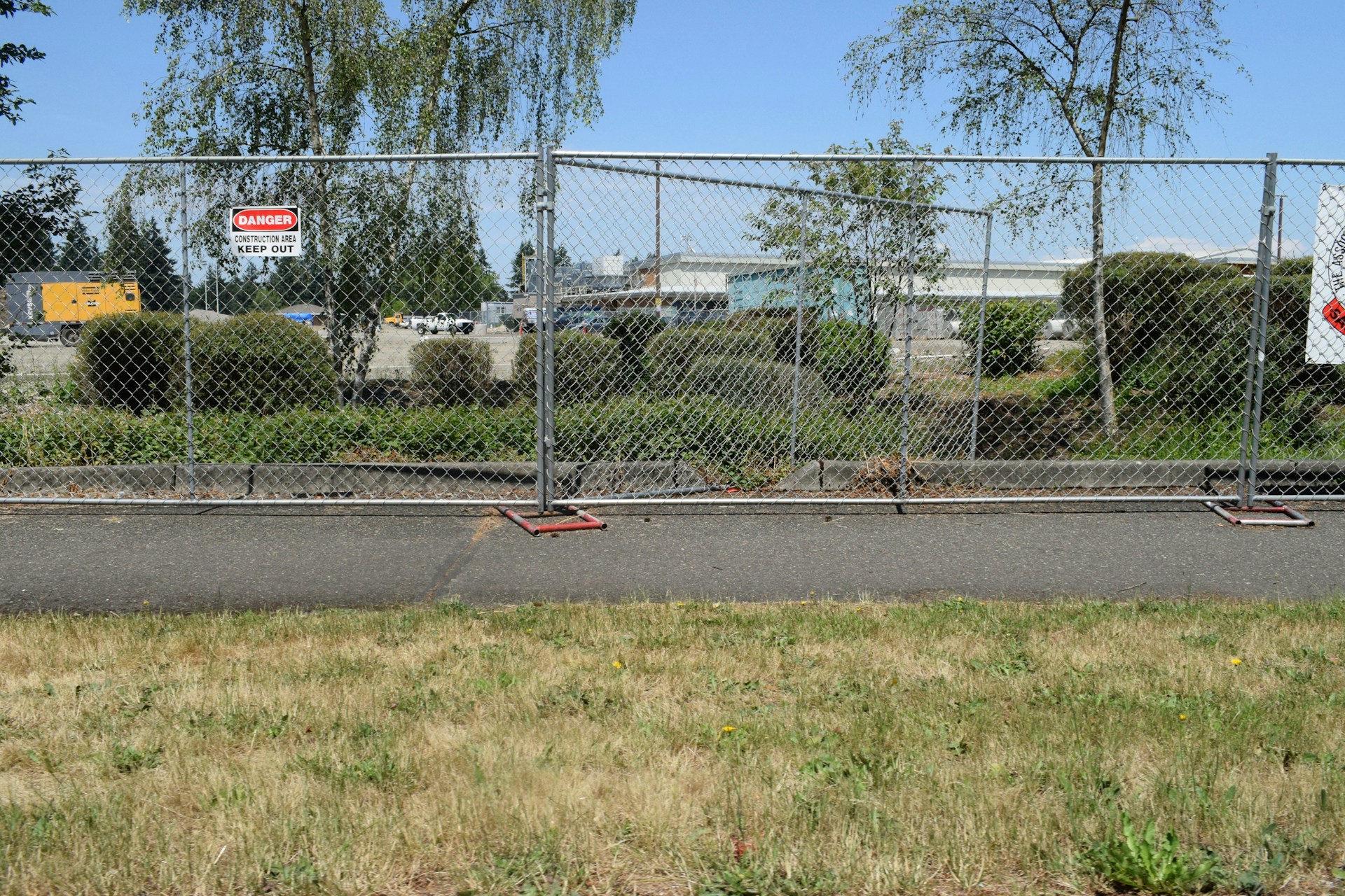 A chain-link fence with signs warning of a construction site surrounds an area with bushes and trees. Behind the fence, construction equipment and vehicles are visible, indicating work in progress. The scene also includes a paved pathway and grass in the foreground.