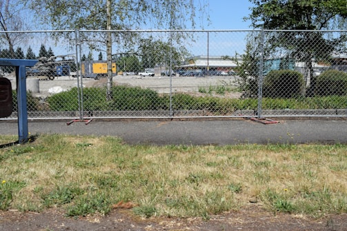 A chain-link fence separates a grassy area from an industrial or construction site. In the background, there are scattered vehicles and machinery, as well as buildings and trees. The sky is clear and blue, indicating a sunny day.