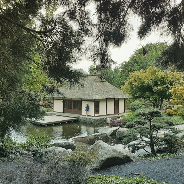 A traditional Japanese-style building with a thatched roof sits peacefully next to a serene pond. Surrounding the structure is lush greenery, with a variety of trees framing the scene. A person stands on a wooden deck extending over the water, adding a human element to the tranquil setting. Rocks and landscaped gardens fill the foreground, enhancing the natural beauty.