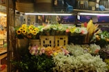 A vibrant flower shop display featuring sunflowers, mixed bouquets, and various types of colorful flowers arranged in baskets and pots. The display is well-lit and surrounded by baskets and other market items.
