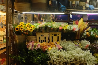 A vibrant flower shop display featuring sunflowers, mixed bouquets, and various types of colorful flowers arranged in baskets and pots. The display is well-lit and surrounded by baskets and other market items.