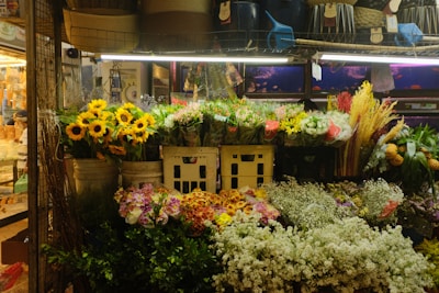 A vibrant flower shop display featuring sunflowers, mixed bouquets, and various types of colorful flowers arranged in baskets and pots. The display is well-lit and surrounded by baskets and other market items.
