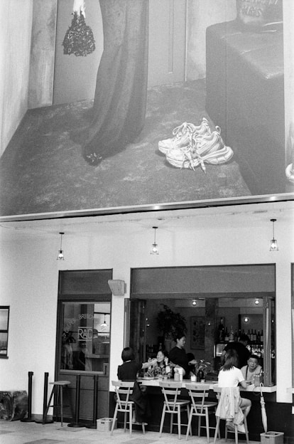 A black and white image featuring an outdoor café scene with people seated at tables, engaged in conversation and dining. Above, there is a large advertisement showcasing a standing figure in a long dress holding a handbag with a pair of sneakers placed on the ground.