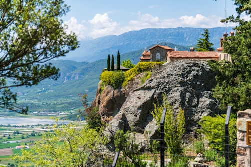 A monastery is perched atop a rocky cliff surrounded by dense green vegetation, with mountains and a clear blue sky in the background. The monastery has terracotta roof tiles and light-colored walls. Below, expansive farmlands stretch into the distance.