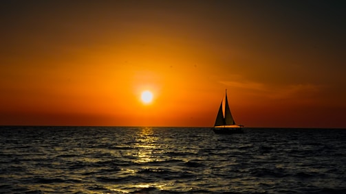 Sunset view of a sailboat silhouette against a sky painted with warm orange and pink hues over calm seas.