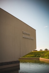 A large, modern building with minimalistic architecture stands next to a body of water. The building is labeled 'Academic Hall' and is positioned against a clear sky. The environment is serene with palm trees visible in the background across a grassy hill.