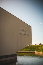 A large, modern building with minimalistic architecture stands next to a body of water. The building is labeled 'Academic Hall' and is positioned against a clear sky. The environment is serene with palm trees visible in the background across a grassy hill.