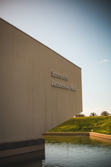 A large, modern building with minimalistic architecture stands next to a body of water. The building is labeled 'Academic Hall' and is positioned against a clear sky. The environment is serene with palm trees visible in the background across a grassy hill.