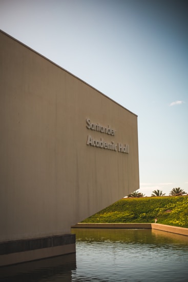 A large, modern building with minimalistic architecture stands next to a body of water. The building is labeled 'Academic Hall' and is positioned against a clear sky. The environment is serene with palm trees visible in the background across a grassy hill.