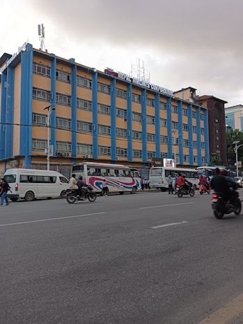 A multi-story building with a sign reading 'Nepal Airlines Corporation' is visible. The structure features a blue and beige color scheme and includes numerous windows on each floor. In front of the building, there are several vehicles, including buses and motorbikes, with people walking or riding along the street. The weather appears overcast, suggesting a cloudy day.