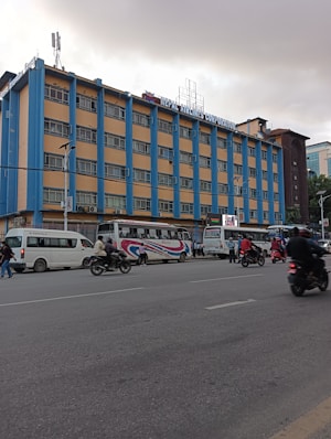 A multi-story building with a sign reading 'Nepal Airlines Corporation' is visible. The structure features a blue and beige color scheme and includes numerous windows on each floor. In front of the building, there are several vehicles, including buses and motorbikes, with people walking or riding along the street. The weather appears overcast, suggesting a cloudy day.