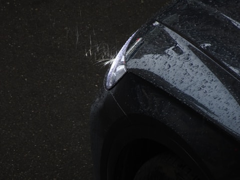 A close-up view of a car's headlight with water droplets scattered across the surface of the dark vehicle. The droplets are visible on the dark, glossy hood and are illuminated by natural light.