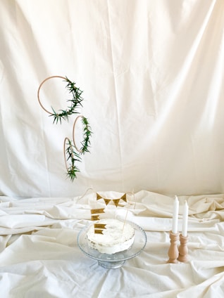 A thoughtfully arranged cake on a glass stand with simple decorations. The cake has a rustic look with minimal icing and is adorned with small paper flags bearing the words 'ONE' and 'Happy Birthday.' To the right, three white candles are held in a wooden candle holder. The background is a plain, cream-colored fabric, creating a minimalist and elegant atmosphere. Greenery is artfully entwined around circular frames suspended beside the cake.