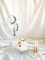 A thoughtfully arranged cake on a glass stand with simple decorations. The cake has a rustic look with minimal icing and is adorned with small paper flags bearing the words 'ONE' and 'Happy Birthday.' To the right, three white candles are held in a wooden candle holder. The background is a plain, cream-colored fabric, creating a minimalist and elegant atmosphere. Greenery is artfully entwined around circular frames suspended beside the cake.