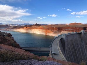 A large concrete dam spans across a river, with a curved structure and a road on top. The body of water behind the dam is surrounded by red and orange layered rock formations, typical of a canyon landscape. The sky above is wide and open, with scattered white clouds and blue hues.
