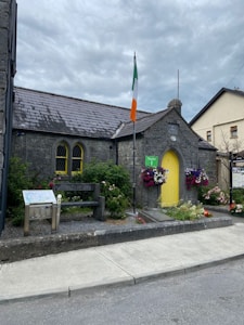A small, stone building with a sloped roof and a yellow door is adorned with vibrant flowers. An Irish flag is prominently displayed in front. There's a wooden bench and an informational sign nearby. The setting is picturesque with a cloudy sky overhead.