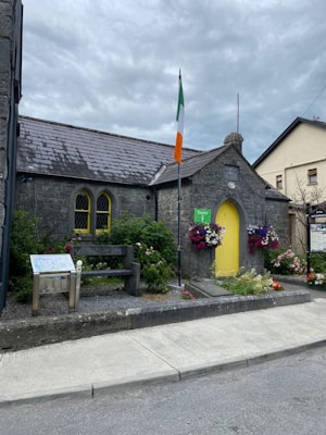 A small, stone building with a sloped roof and a yellow door is adorned with vibrant flowers. An Irish flag is prominently displayed in front. There's a wooden bench and an informational sign nearby. The setting is picturesque with a cloudy sky overhead.