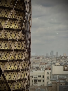 A modern building with a distinctive diamond-shaped glass facade on the left side, set against the backdrop of a cityscape with numerous older residential buildings. The sky is overcast, giving a muted ambiance to the scene.