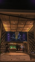 A modern entrance features an intricately designed ceiling with wooden panels and embedded lighting. The wall art integrates abstract shapes and figures illuminated with blue and green hues. Below, a reception area is visible with a desk and a person behind it, creating an inviting atmosphere.