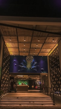 A modern entrance features an intricately designed ceiling with wooden panels and embedded lighting. The wall art integrates abstract shapes and figures illuminated with blue and green hues. Below, a reception area is visible with a desk and a person behind it, creating an inviting atmosphere.