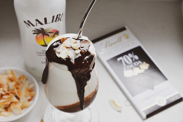 A dessert beverage garnished with coconut flakes is elegantly presented in a glass, topped with a generous drizzle of chocolate syrup. A spoon is inserted into the drink. Adjacent to the glass, a bottle of Malibu coconut rum and a bar of Lindt 70% dark chocolate are visible. A small dish containing toasted coconut flakes completes the presentation.