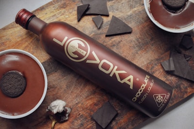A bottle labeled as chocolate fusion liqueur rests on a wooden surface, surrounded by chocolate-themed items. There are two bowls with rich, dark chocolate pudding topped with cookies, alongside scattered pieces of dark chocolate.