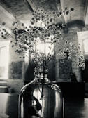 A glass vase holding delicate flowers with small, clustered blooms is placed on a table. The backdrop features a rustic interior with exposed brick walls and arched ceilings. Light streams in through the windows, creating a serene and vintage atmosphere.