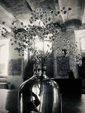 A glass vase holding delicate flowers with small, clustered blooms is placed on a table. The backdrop features a rustic interior with exposed brick walls and arched ceilings. Light streams in through the windows, creating a serene and vintage atmosphere.